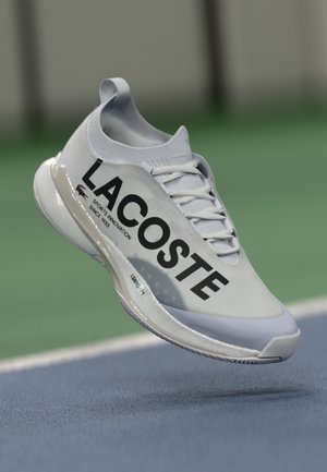 White sports shoe made of synthetic material featuring a rounded toe, laced front, and large black "LACOSTE" branding on the side.