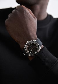 Reloj deportivo negro con correa de goma negra texturizada, hardware plateado, detalles de cronógrafo y una esfera negra con acentos en blanco y rojo.