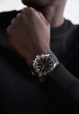 Schwarze Sportuhr mit einem strukturierten schwarzen Gummiarmband, silbernen Beschlägen, Chronographendetails und einem schwarzen Zifferblatt mit weißen und roten Akzenten.