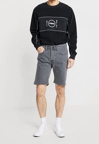 Uomo che indossa un dolcevita nero a maniche lunghe con testo bianco e pantaloncini di jeans grigi, calze bianche e scarpe da ginnastica bianche, in piedi con una mano in tasca.