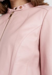 Ljusrosa läderjacka med dragkedja framtill, rund krage och tryckknapsdetaljer. Har en slät textur och subtila sömdetaljer.