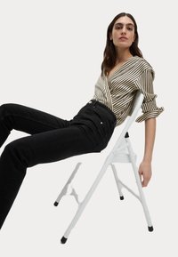 Gestreepte zwart en crèmekleurige wikkelblouse met opgerolde mouwen, gecombineerd met hoge taille zwarte jeans. Het model zit op een witte klapstoel.