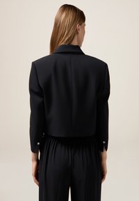 Blazer nero corto con un design strutturato, dotato di colletto a punta e dettagli con bottoni sulle maniche. Tessuto a trama liscia.