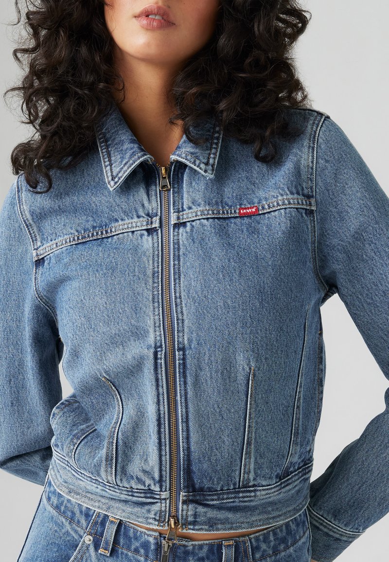 Chaqueta de mezclilla azul claro con cierre de cremallera en el frente, cuello puntiagudo y dos bolsillos frontales. Presenta costuras visibles y una pequeña etiqueta de logo roja.