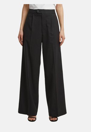 Femme portant un pantalon large à rayures noires, taille haute et bouton sur le devant, associé à des sandales noires à bouts ouverts et un top blanc raccourci.