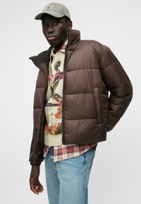 Brązowa kurtka puchowa z wysokim kołnierzem i kieszeniami na zamek, nałożona na wzorzysty sweter, w połączeniu z jasnoniebieskimi dżinsami i czapką.