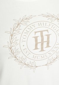 White cotton t-shirt featuring a beige circular logo with "TOMMY HILFIGER" and "EST. MCMLXXXV," surrounded by a leaf design.