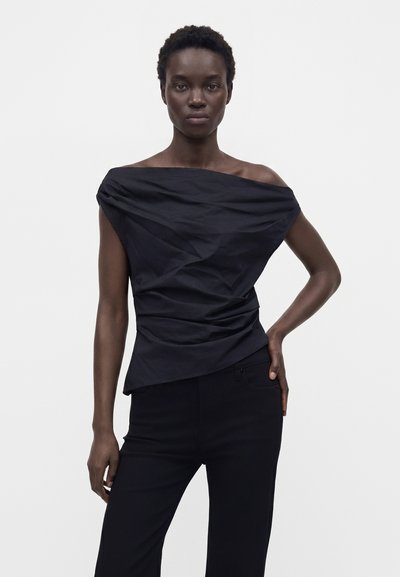 Femme aux cheveux courts et bouclés portant un haut noir asymétrique épaules dénudées et un pantalon noir, posant avec une main sur la hanche devant un fond uni.