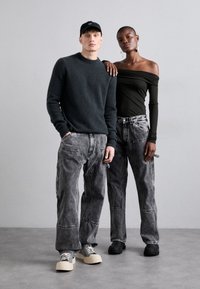 To modeller står sammen. Den mandlige model har en mørkegrå sweater og falmede grå cargo jeans på. Den feminine model har en sort off-the-shoulder top og grå jeans på.