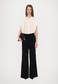 Bala balta caurspīdīga blūze ar plisētiem elementiem un pogu aizdari, saskaņota ar augsto jostasvietu melniem platām biksēm. Modelis tur melnu clutch somu.