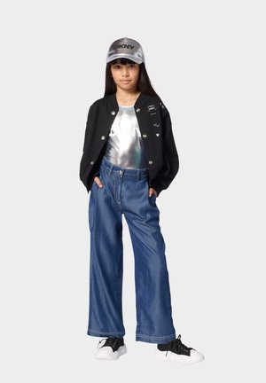 Jeune fille portant une casquette argentée DKNY, un haut métallique argenté, une veste noire boutonnée, un jean bleu ample et des baskets noires et blanches, debout les mains dans les poches.