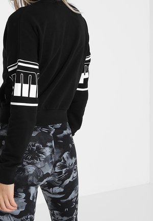 Kvinde iført sort croppet sweatshirt med hvide grafiske striber på ærmerne og sorte og grå blomstrede leggings, stående mod hvid baggrund.