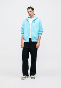 Ανοιχτό μπλε φούτερ με φερμουάρ και μπροστινή τσέπη, φορεμένο πάνω από μια λευκή μπλούζα, συνδυασμένο με μαύρο παντελόνι και πολύχρωπα αθλητικά παπούτσια.