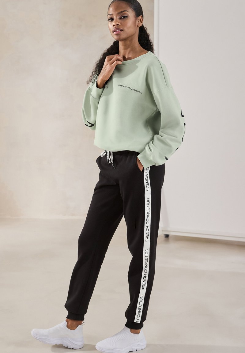 Mintgrüner Sweatshirt mit schwarzen Akzenten, kombiniert mit schwarzen Jogginghosen, die weiße seitliche Streifen haben. Weiße Sport-Sneaker runden den Look ab.