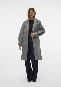 Manteau en laine gris avec un col châle, deux boutons et une coupe décontractée. Porté sur un haut noir et associé à un jean sombre et des chaussures noires.