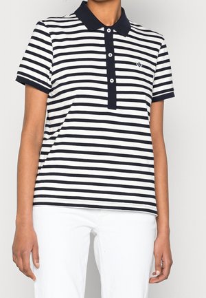 Persoon draagt een zwart-wit horizontaal gestreept poloshirt met korte mouwen, een zwarte kraag en een witte broek.