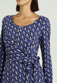 Sukienka z długim rękawem z geometrycznym wzorem w kolorach niebieskim, czarnym i białym. Z taliowanym paskiem, dekoltem w kształcie łódki oraz gładką fakturą materiału.