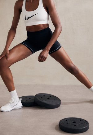 Atlet iført hvid sports-bh og sorte shorts strækker benene ved siden af sorte vægtskiver på fitnessgulvet.