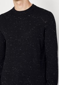 Schwarzer Strickpullover mit melierter Textur, runder Ausschnitt und langen Ärmeln. Das Material zeigt subtile Farb- und Musterabweichungen.