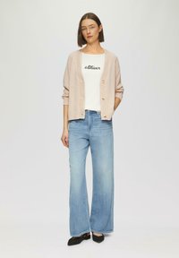 Gilet tricoté rose clair, t-shirt blanc avec texte noir, jeans larges bleu clair et chaussures noires. Tenue simple et décontractée.