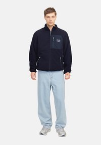 Chaqueta de forro polar en color marino con bolsillos con cremallera, panel en el pecho de color contrastante, detalle del logo en blanco, combinada con jeans de color azul claro y zapatillas grises.