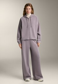 Set di abbigliamento casual grigio composto da una felpa con zip e pantaloni a gamba larga, realizzato in tessuto morbido, con una vestibilità rilassata e dettagli con cordini.