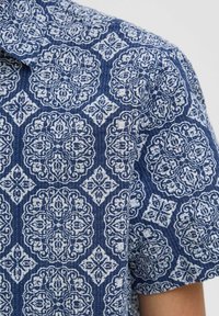 T-shirt trumpu rankovu jūros mėlynos spalvos, puoštas baltu sudėtingu gėlių ir geometriniu raštu su subtilia tekstūra ir detalėmis.