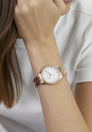 Roségouden horloge met een witte wijzerplaat, metalen armband en decoratieve kristalaccenten rond de lunette. Gladde afwerking en eenvoudige uurmarkeringen.