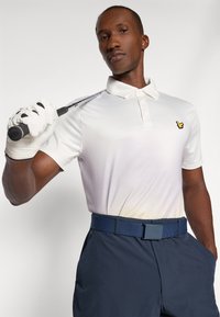 White short-sleeve polo shirt with a yellow logo, paired with navy pants and a ribbed blue belt, holding a golf club with a white glove.