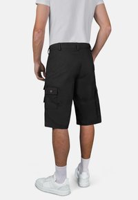 Schwarze Cargo-Shorts mit knöpfbaren Seitentaschen, lockerer Passform und glatter Textur. Kombiniert mit weißen Sneakers und Socken.