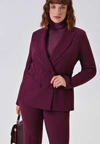 Frau in einem maßgeschneiderten, lilafarbenen, doppelt geknöpften Blazer, passenden Hosen und einem glitzernden, lilafarbenen Rollkragenpullover, die eine dunkel strukturierte Handtasche hält.