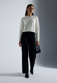 Maglione bianco a maglia grossa con orlo a coste, abbinato a pantaloni neri a vita alta. Una borsa mini nera e tacchi neri a punta completano il look.