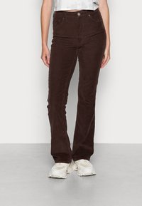 Calças de corduroy castanhas, com corte flare e cintura alta, apresentando bolsos frontais e fecho com botão. O material tem um acabamento suave e texturizado.