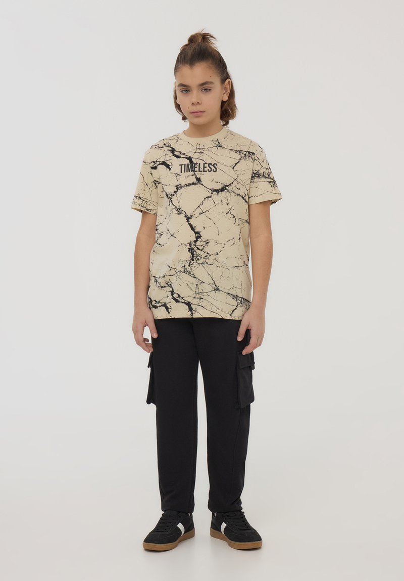 T-shirt beige con motivo di marmorizzazione nera e testo "TIMELESS", abbinato a pantaloni cargo neri e sneakers nere con suole in gomma.