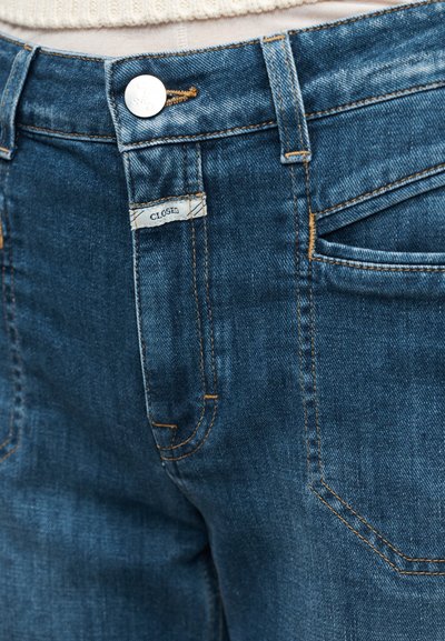Κοντινή άποψη μπλε τζιν τζιν με μεταλλικό κουμπί, μπροστινή τσέπη, θηλιές ζώνης και μια μικρή υφασμάτινη ετικέτα "CLOSED" κοντά στο φερμουάρ.