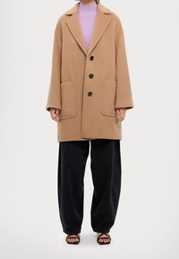 Abrigo de lana marrón claro con solapa, dos grandes bolsillos frontales y tres botones, llevado sobre una blusa de punto morada y pantalones de pierna ancha negros.