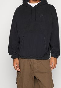 Person som bär en svart Nike fleecehuva med snoddar och en framtida dragkedjeficka, ihop med bruna cargobyxor.