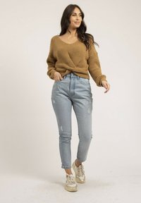 Pull en tricot marron avec un devant torsadé, associé à un jean déchiré bleu clair et des baskets avec des accents dorés. Fond minimaliste.