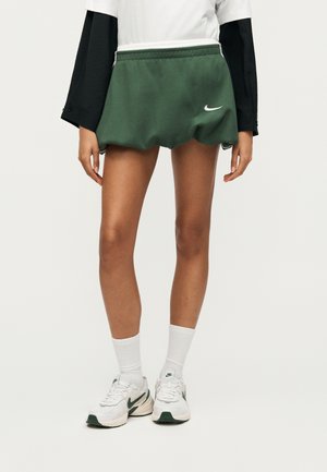 Το μοντέλο φοράει πράσινα σορτς Nike, λευκές καλτσούλες μέχρι τον αστράγαλο, λευκά και πράσινα αθλητικά Nike και μαύρο-λευκό μακρυμάνικο τοπ με στρώσεις, όρθιο.