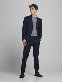 Marineblauer Anzug mit einem taillierten Sakko und passender Hose, getragen über einem hellgrauen Hemd mit Kragen. Dunkle Schuhe runden das Ensemble ab.