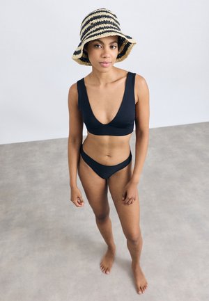 Μαύρο μπικίνι με βαθύ ντεκολτέ και χαμηλό παντελόνι. Φορεμένο με καπέλο μπαούλα με μαύρες και μπεζ ρίγες. Απλή φόντο.