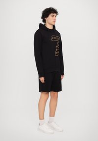 Black zip-up hoodie with gold text, paired with black shorts. Model wears white sneakers with light-colored socks. Smooth fabric texture.