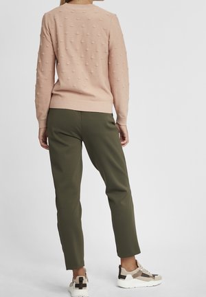 Vrouw die met haar rug naar de camera staat, gekleed in een textuur lichtroze trui, olijfgroene broek en beige en witte sneakers op een effen achtergrond.