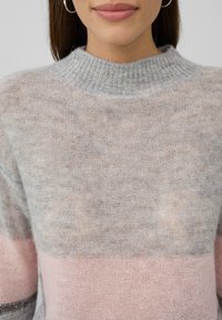 Ženska v mehkem pletenem puloverju z barvnimi bloki v sivi, roza in beli barvi, z robom visoke ovratnice in vidnimi srebrnimi obročastimi uhani.