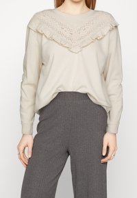 Bluza w jasnobeżowym kolorze z koronkową nakładką i marszczonym dekoltem. Połączona z szarymi prążkowanymi spodniami o luźnym kroju i miękkiej teksturze.