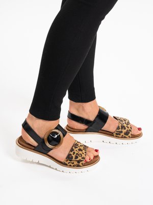 Pies con sandalias de cuero negro y estampado de leopardo, con una gran hebilla de metal y suelas gruesas blancas, combinadas con leggings negros.