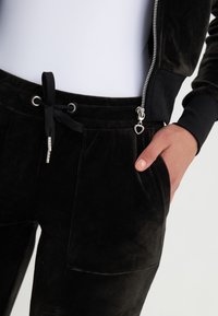 Chándal de terciopelo negro que incluye una chaqueta con cremallera, cinturilla con cordón y bolsillos laterales con un pequeño acento en forma de corazón.