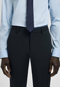 Granatowe spodnie o płaskim przodzie, noszone z jasnoniebieską koszulą i granatowym krawatem. Gładka tkanina; minimalistyczny design; standardowe dopasowanie z bocznymi kieszeniami.