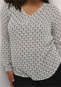 Femme portant un haut à manches longues noir et blanc à motifs en forme de goutte d'eau verticale, associé à un pantalon noir, se tenant devant un fond blanc.