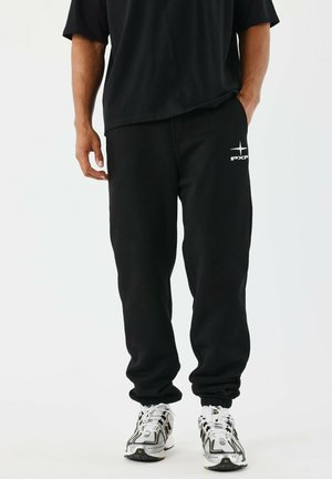 Personne portant un pantalon de survêtement noir avec un logo blanc sur la cuisse et des baskets blanches et noires, se tenant debout avec une main dans la poche.